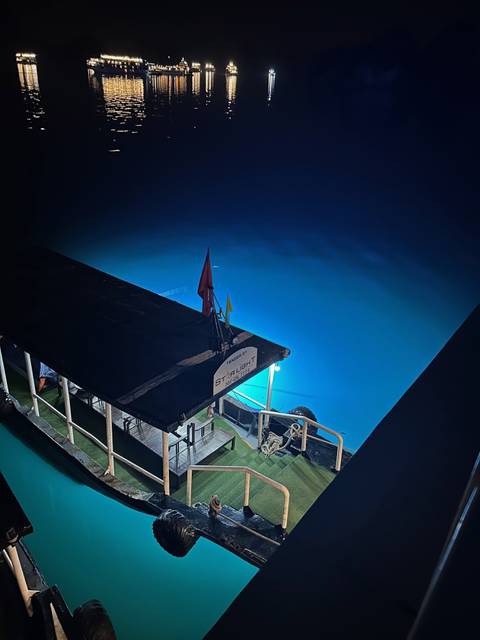       Small cruise launch illuminated against glowing blue water at night.
  