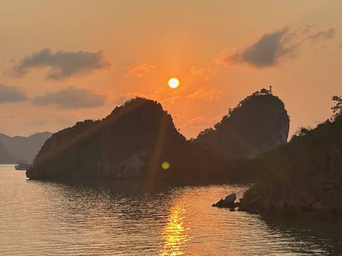      Golden-hour sunset over karst islets on tranquil bay waters, sun low behind a jagged peak.
  