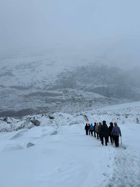       Line of hikers in colorful jackets walking up a snowy glacier with steep, mist-covered mountain walls ahead.
  