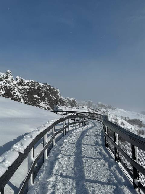       Snow-covered boardwalk curving alongside rugged cliffs under clear blue sky in a national park.
  