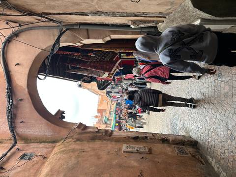       Busy covered archway opening onto a bustling market street with shoppers and rugs hanging.
  