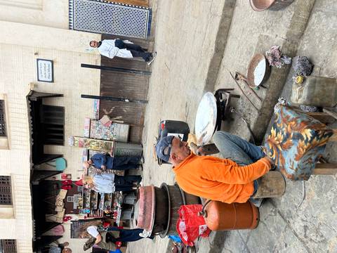       Metalworker seated on a street corner shaping cookware while bystanders watch.
  