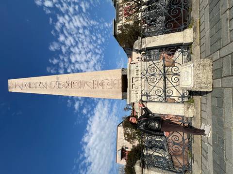       A traveler poses beside the carved base of a towering obelisk under blue skies.
  