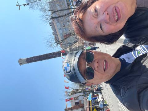       A couple’s selfie partially frames an ancient column in a lively urban square.
  