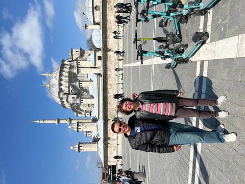       Two travelers stand before an Ottoman mosque with electric scooters nearby.
  