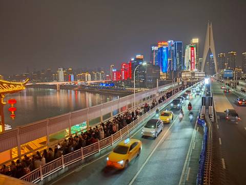       Colorful illuminated skyscrapers reflect on a river beside a crowded pedestrian bridge filled with evening commuters in Chongqing.
  