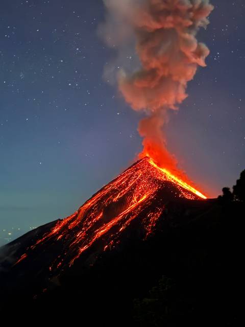       Dramatic night eruption of a cone-shaped volcano with glowing lava rivers and ash plume against a twilight sky.
  