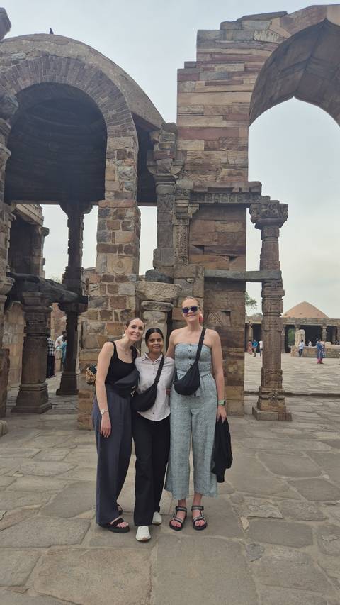       Friends smiling among ancient stone pillars and arches at the Qutub archaeological complex.
  