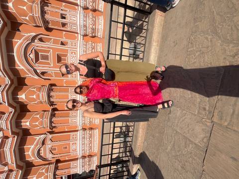       Three women pose together in front of the ornate orange-and-white facade of Jaipur’s Hawa Mahal under warm sunlight.
  