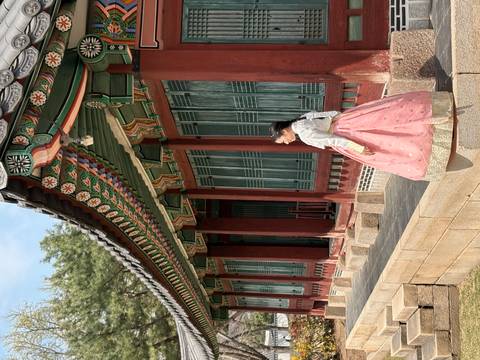       Woman in traditional pink hanbok strolls along the colonnade of a historic Korean palace building.
  