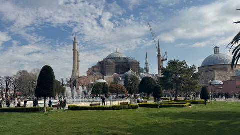       Wide garden view of Hagia Sophia with fountains, domes and cranes under patchy clouds.
  