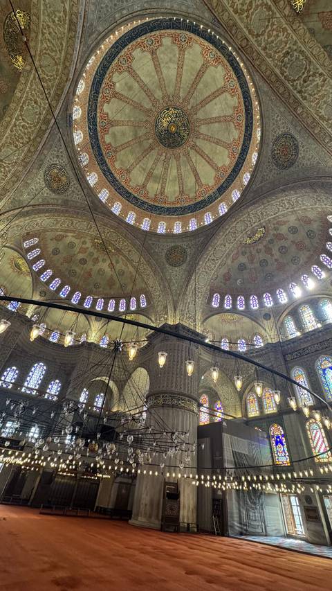       Intricate interior domes and stained glass of the Blue Mosque lit by hanging lamps.
  