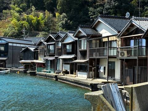      Row of traditional wooden boathouses lining calm blue water in rural Japan
  