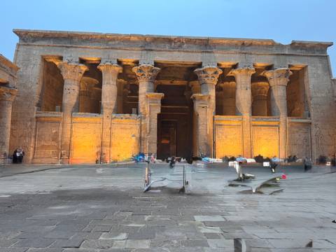       Facade of an ancient Egyptian temple lit warmly at dusk with blurred visitors walking across the courtyard
  