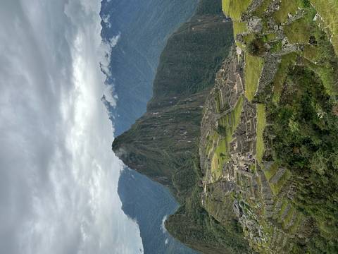       Wide view of Machu Picchu citadel nestled below a steep green peak under overcast skies.
  