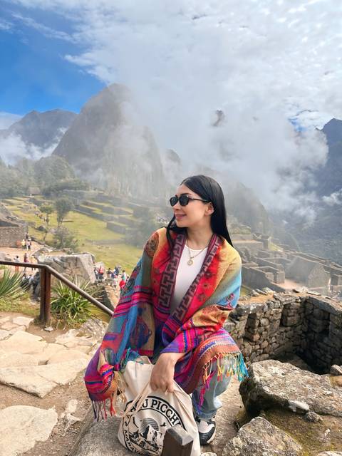       A woman wrapped in a colorful shawl looks over misty Machu Picchu ruins partially veiled by clouds.
  