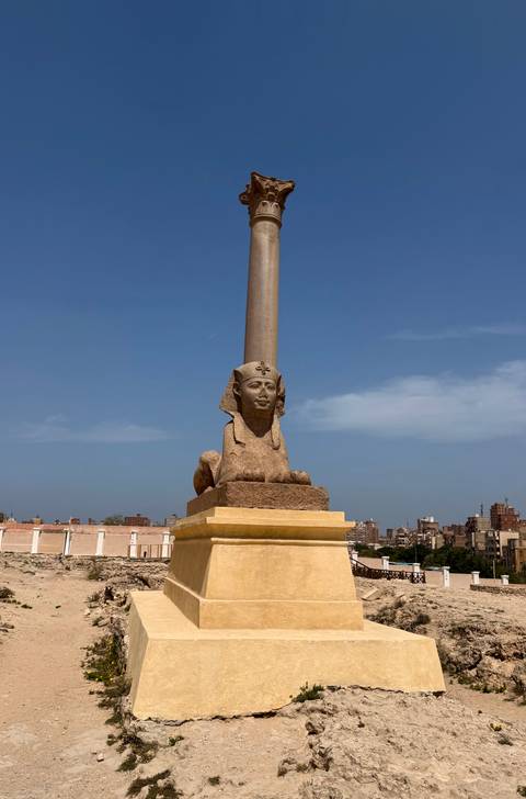       Sphinx-headed statue supports an ancient column against a blue Alexandria sky.
  
