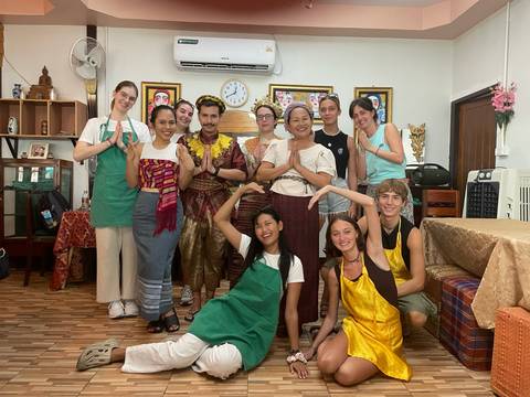       Cooking class participants pose inside a Thai kitchen wearing aprons and traditional sashes.
  
