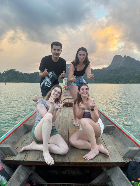       Friends make heart gestures while sitting on the prow of a long-tail boat on a lake at sunset.
  