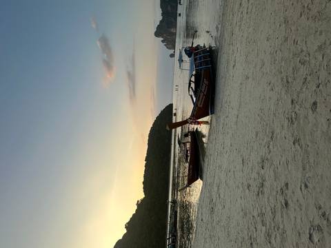       Traditional long-tail boats rest on sandy shore at dusk with forested hills beyond.
  