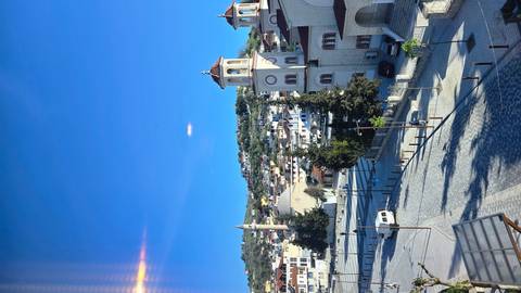       Elevated view over a quiet Albanian town square with mosques and hillside houses under a deep blue sky.
  