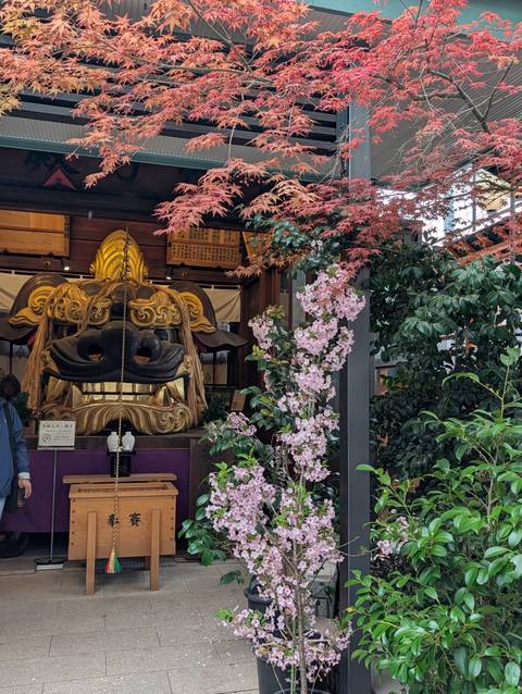       Decorative lion mask displayed beside blooming cherry blossoms and maple leaves at a shrine
  