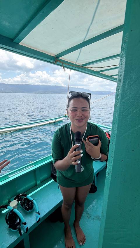       Smiling woman on a boat holding a microphone and phone with turquoise sea and islands in the background
  