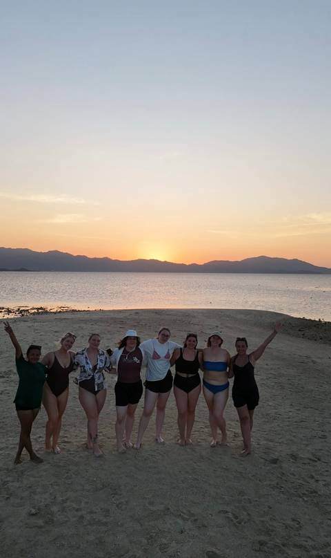       Group of travelers silhouetted against a pastel sunset on a tropical beach.
  