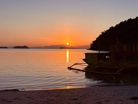       Traditional outrigger boat anchored on calm water reflecting a vibrant orange sunset.
  