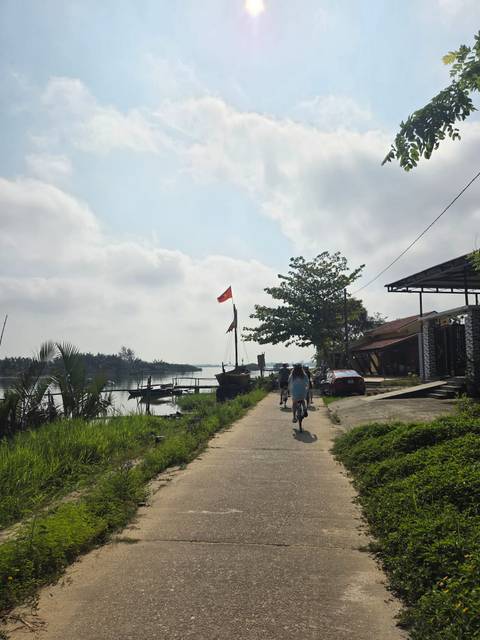       Cyclists riding along a riverside path beneath a Vietnamese flag with boats and lush greenery nearby
  