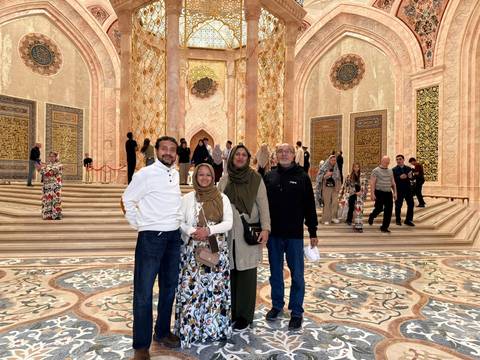       A small group poses on ornate marble steps beneath a grand golden interior dome with Islamic motifs.
  