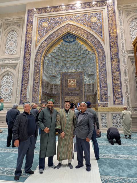       Four men wearing traditional caps stand before an intricately tiled mosque interior.
  