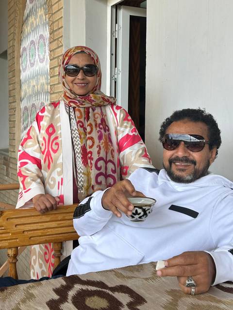       A man wearing sunglasses enjoys a cup of tea while a woman stands beside him on a balcony.
  