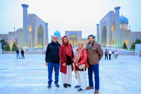       Four travellers stand in front of the illuminated Registan madrasas at dusk in Samarkand.
  