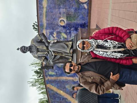      A couple poses before a large bronze statue of astronomer Ulugbek against a blue tiled mural.
  