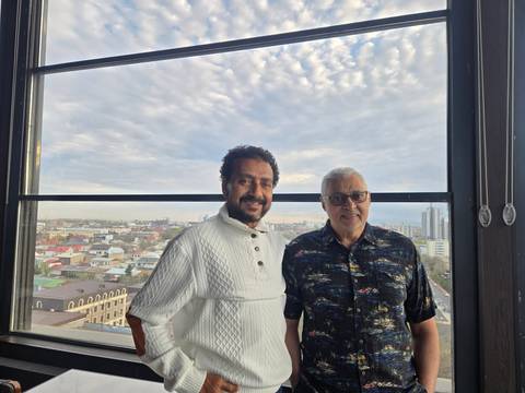       Two men pose by a large window overlooking a sprawling cityscape at dusk.
  
