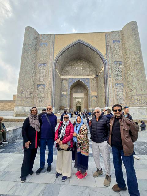       A tour group poses before the grand tiled portal of the Ulugbek Madrasa packed with visitors.
  