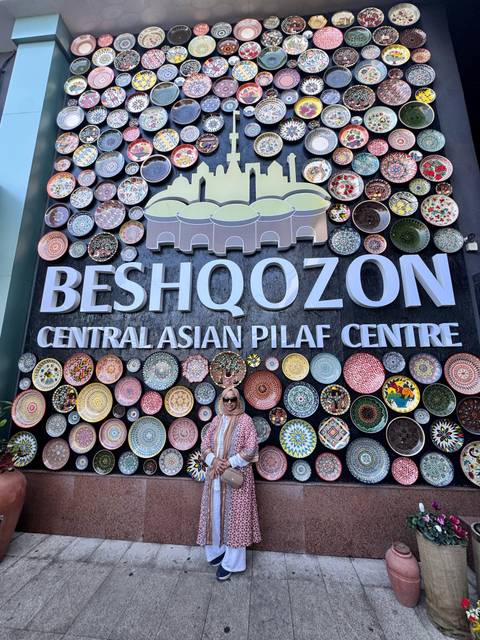       A woman stands beneath a large sign for the Central Asian Pilaf Centre decorated with colorful plates.
  