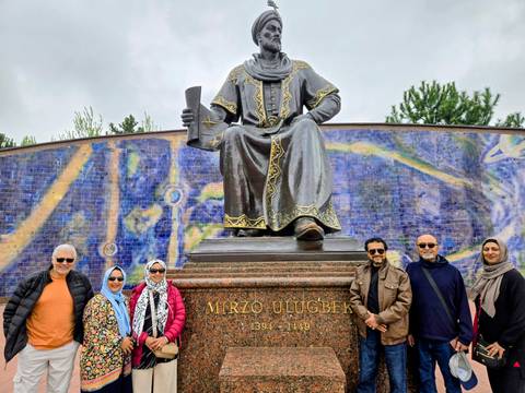       Travellers pose in front of the seated bronze statue of Ulugbek with a colorful mural backdrop.
  