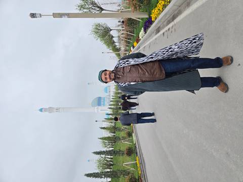       A man in traditional coat stands on a broad avenue leading to a modern white and blue mosque.
  