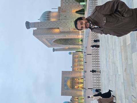       A smiling traveller stands at dusk before the softly lit arches of Registan Square.
  