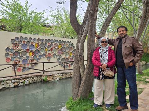       A couple poses beside a decorative wall of colorful ceramic plates next to a narrow canal and trees.
  