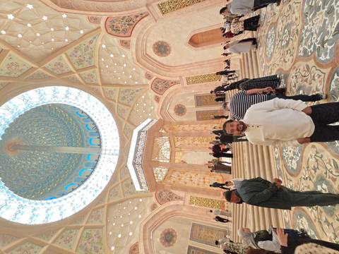       A traveller stands beneath a towering, ornate dome skylight inside an opulent mosque filled with visitors.
  