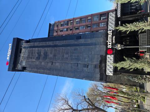       Tall black glass façade of the Azimut Hotel rises against a clear sky with flags at its entrance.
  