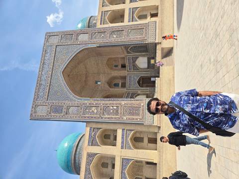       A smiling man stands before a grand tiled portal with turquoise domes under a bright sky.
  
