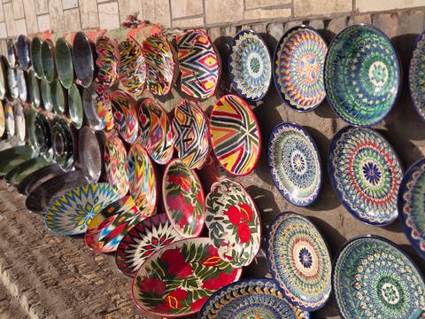       Rows of hand-painted Uzbek ceramic plates and bowls gleam in bright sunlight along a stone wall.
  