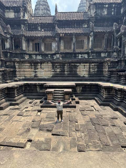       Visitor stands arms wide inside the ancient stone court of Angkor Wat, surrounded by tiered galleries.
  