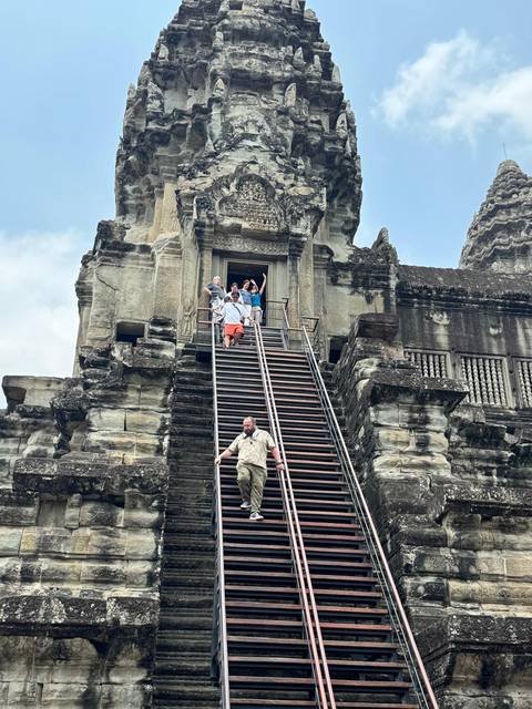       Tourists descend the steep wooden staircase of Angkor Wat’s central tower, waving to the camera.
  