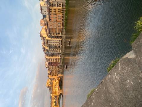       Golden light falls on riverside buildings along the Arno with small boats on the calm water
  