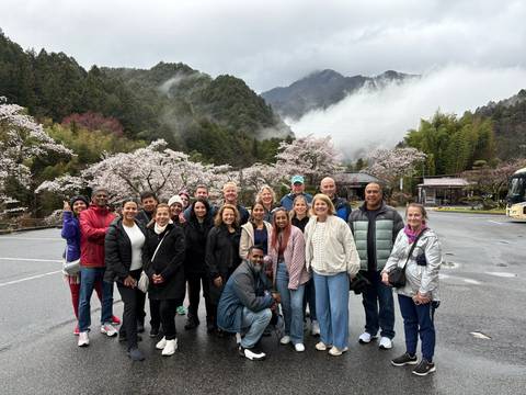       Smiling tour group poses among blooming cherry trees with misty forested mountains rising behind them.
  